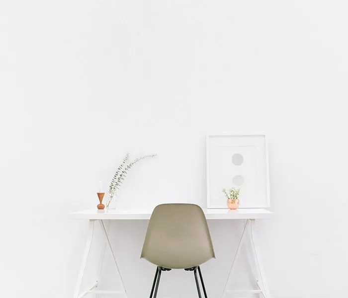 Minimalist workspace with a white desk, green chair, two small potted plants, and a framed print against a white wall.