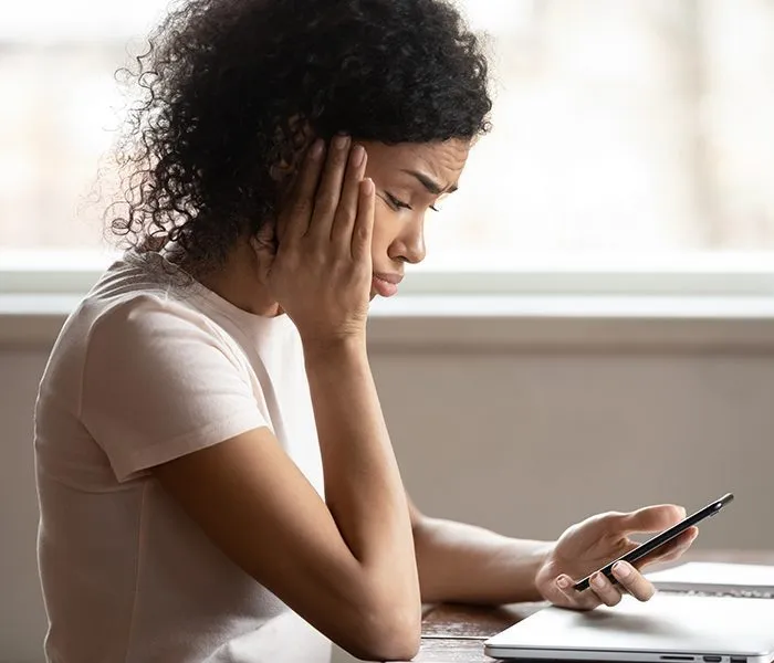 A woman sitting at a table, holding a smartphone in one hand, resting her head on the other, looking at the screen.