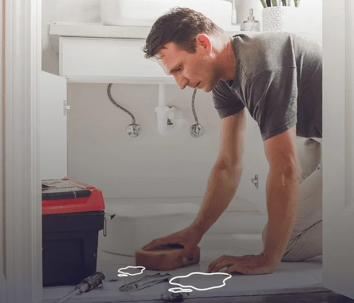 A man kneels on a floor beside a toolbox, inspecting a plumbing issue with water puddles on the ground.