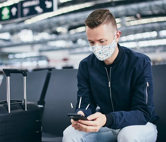 A person with a face mask sitting in an airport, looking at their phone, with a suitcase beside them.