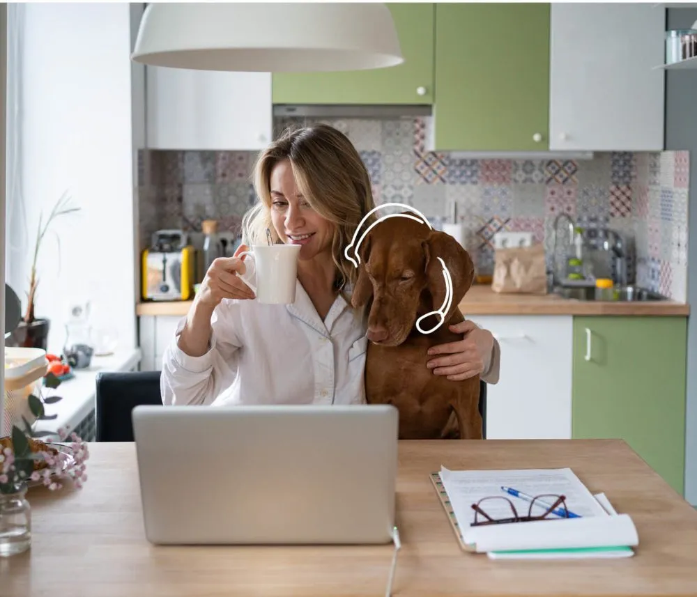 A woman works on a laptop at her kitchen table, holding a mug and hugging a dog wearing headphones.