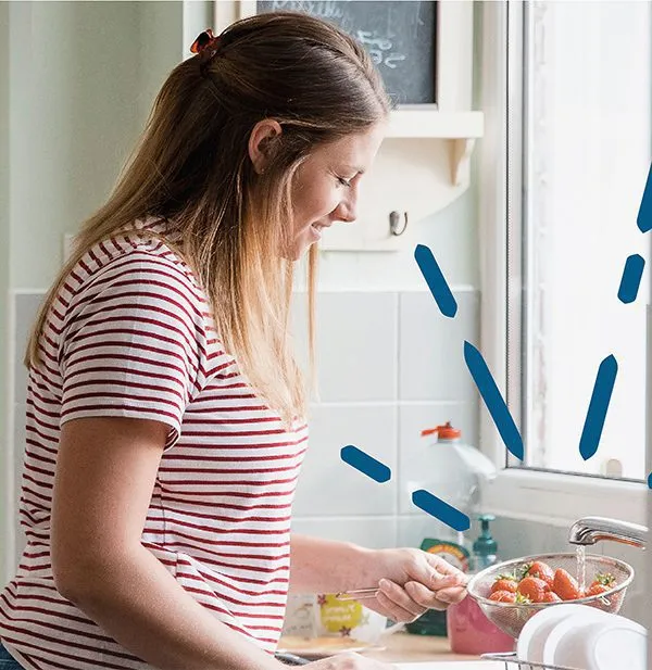 A woman in a striped shirt rinses strawberries in a kitchen sink with natural light coming through the window.