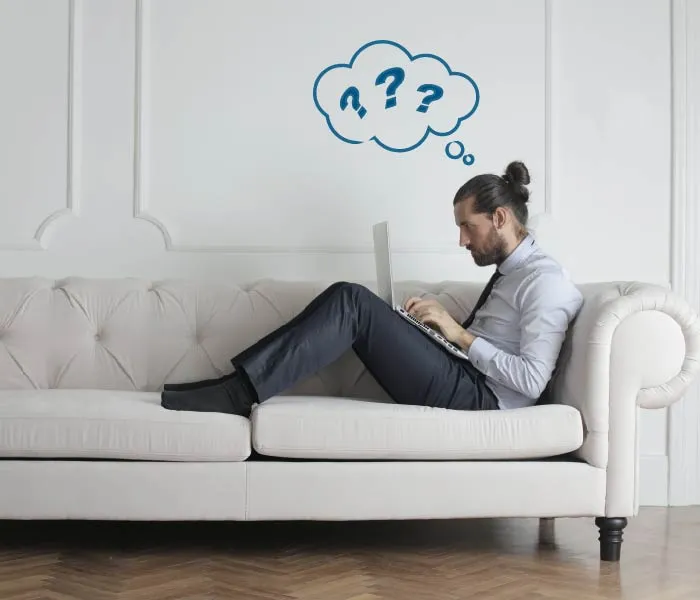 A man with a laptop sits on a couch, appearing deep in thought with a thought bubble containing question marks above his head.