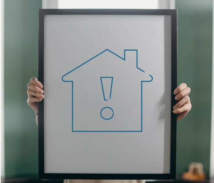 Person holding a framed picture of a house icon with an exclamation mark in the center against a blurred background.