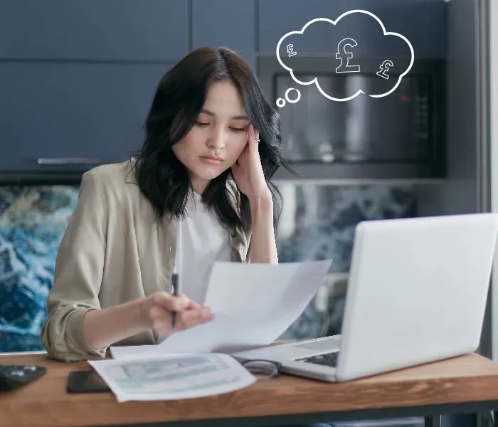 Woman sitting at a desk with a laptop, looking at documents, and a thought bubble with pound symbols above her head.