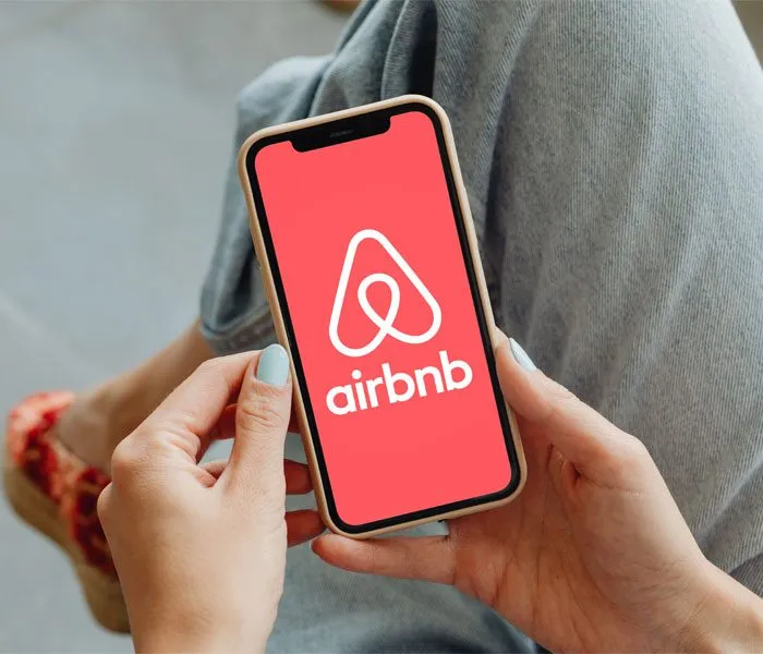 Person holding a smartphone displaying the Airbnb logo on the screen while sitting with legs crossed.