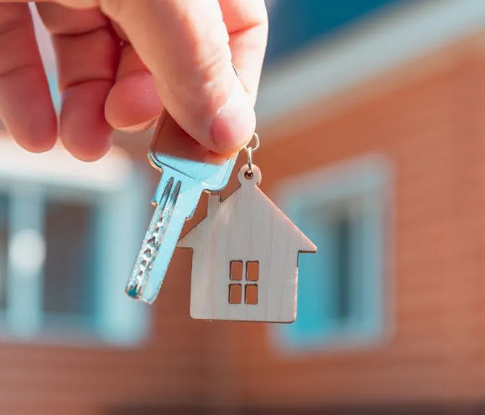 A hand holds a house-shaped keychain and a key in front of a blurred background of a house.