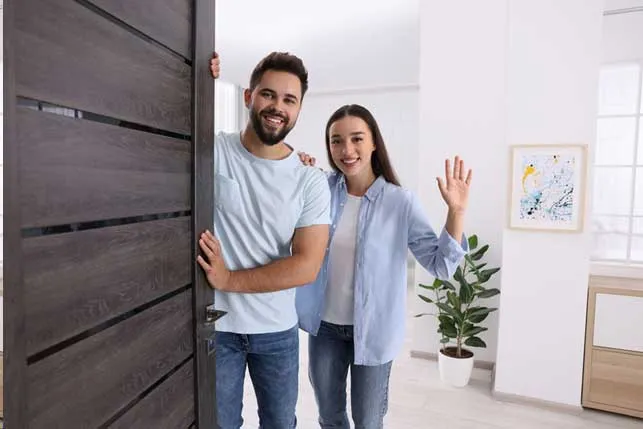 A man and a woman stand at an open door, smiling and waving. They are inside a bright room with a potted plant and a painting.