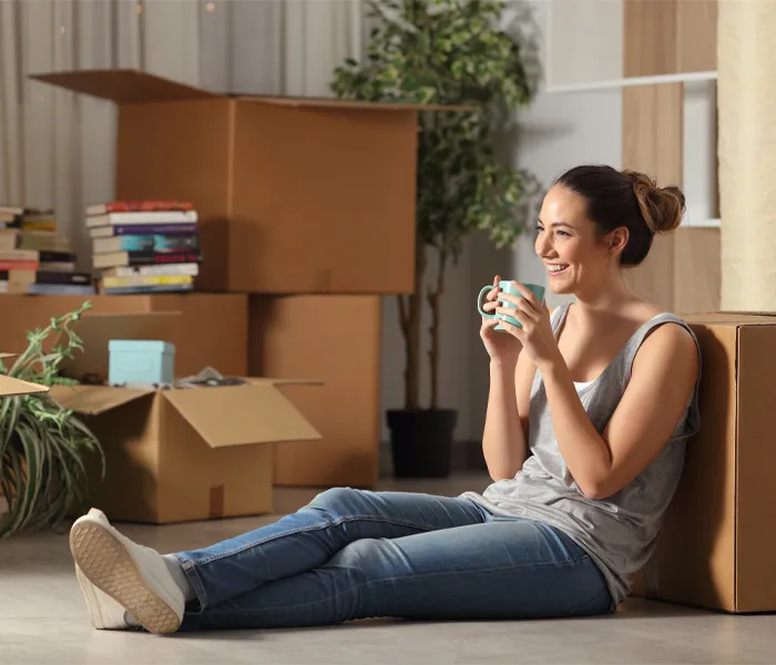 A woman sits on the floor with a mug, surrounded by moving boxes and books, looking content.